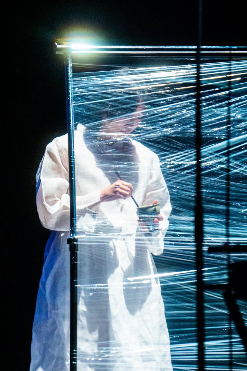 Figure in white robe, partially obscured by a grid of reflective blue-white lines. They hold a small bowl and brush, against a dark background.