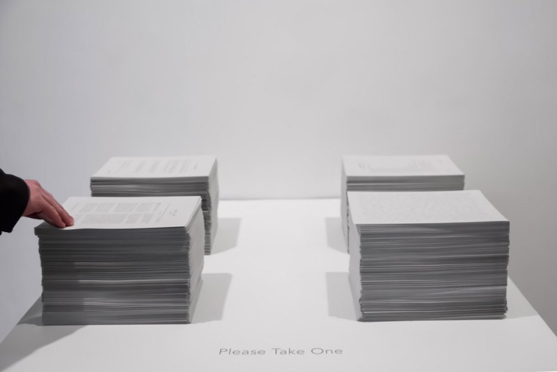 Four stacks of white paper on a white table, a hand reaches for the top sheet of the front left stack. "Please Take One" is written below.