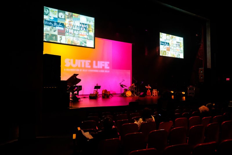 Dark auditorium with a stage featuring musical instruments, bathed in pink and orange light, with two screens displaying collages.