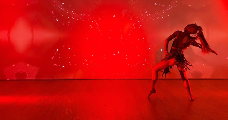 Dancer in a sequined costume with a long ponytail, striking a pose on a wooden stage against a vibrant red background with white speckles.