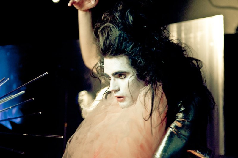 Performer with pale face paint, dark messy hair, and a light pink ruffled collar, looking left.