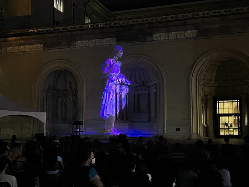 Nighttime projection of a glowing purple female figure onto a building facade, with an audience seated below.