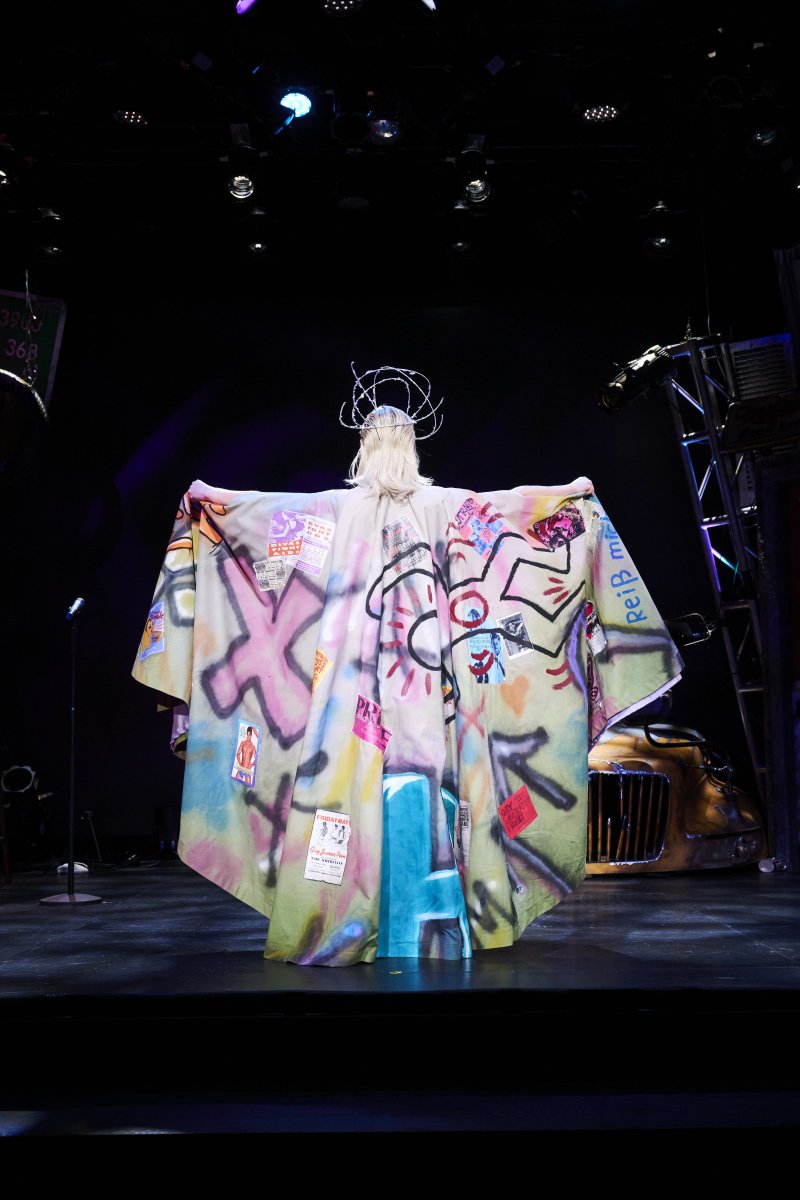 Drag performer in a colorful, graffiti-covered robe and spiky crown, facing away from the viewer on a dark stage with spotlights.