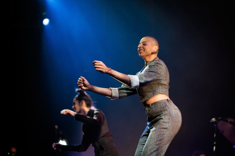 A bald person in a grey suit dances on stage, arms bent, hands open, with another dancer blurred in the background.