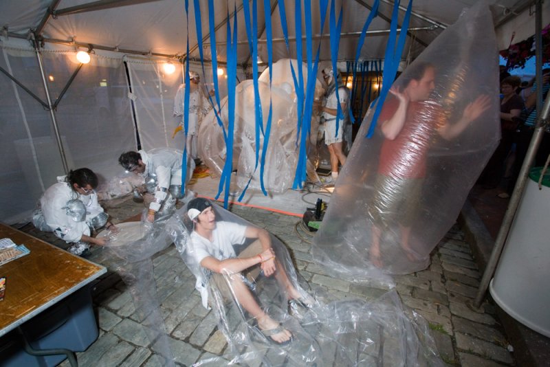 Performance art in a tent with people in clear plastic, blue streamers, and white suits.