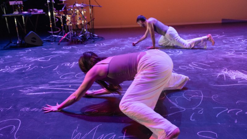 Two dancers in light clothing crawl on a dark stage covered in white chalk writing, under purple and orange lighting.