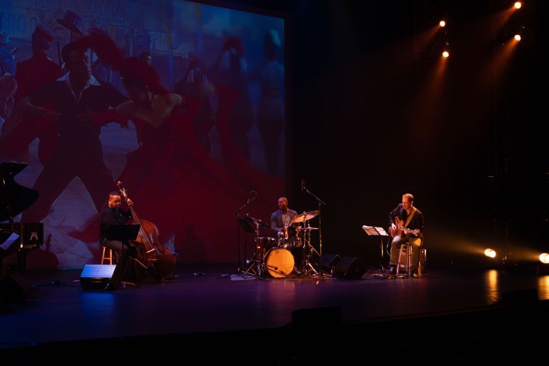 Three musicians on a dark stage, illuminated by spotlights, perform in front of a large screen displaying a projected image of dancers.