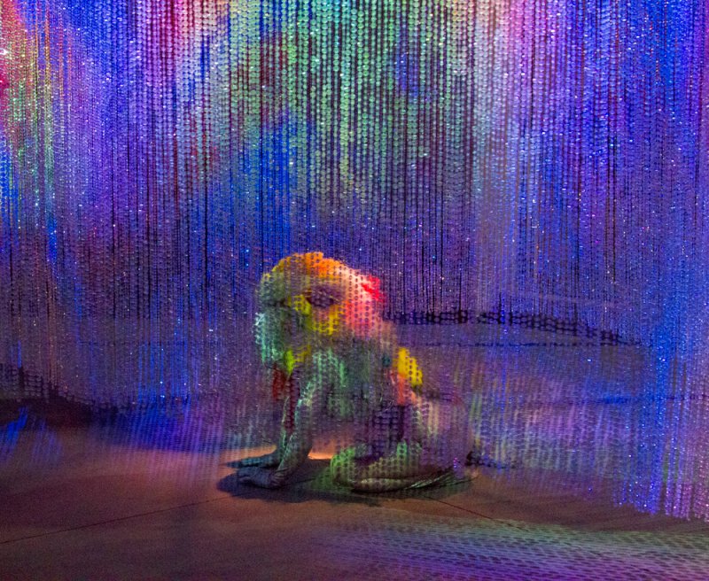 Sculpted dog-like figure splattered with paint sits under a shimmering curtain of colorful lights.