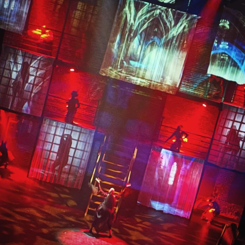 Performance art with red lighting, projections of figures and windows on a grid set, and a figure on stage.