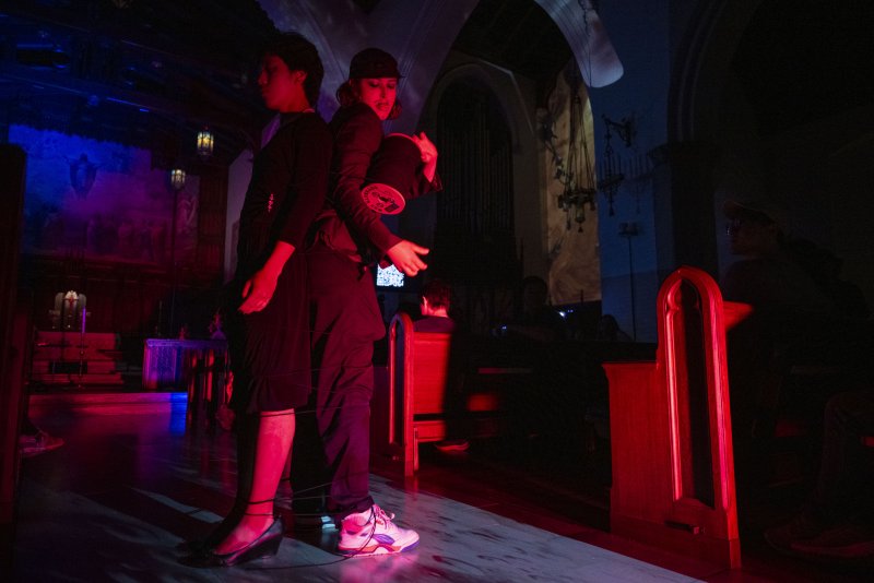 Two performers in a dimly lit church, one in black dress, one in black cap and pants, connected by string, bathed in red and blue light.