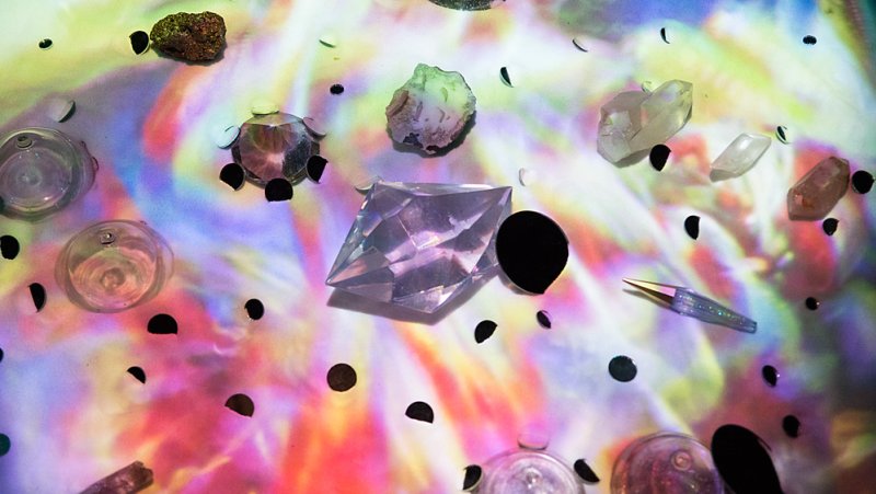 Overhead view of various crystals, rocks, and glass objects on a surface illuminated by shifting colorful lights.