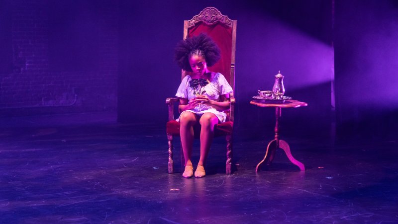 Young woman sits in ornate chair holding black roses, lit in purple. Teapot and cups on small table.