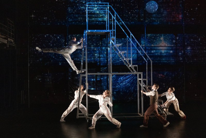 Performers in white and brown costumes interact with a blue metal structure on a dark stage with a starry, brick-patterned backdrop.