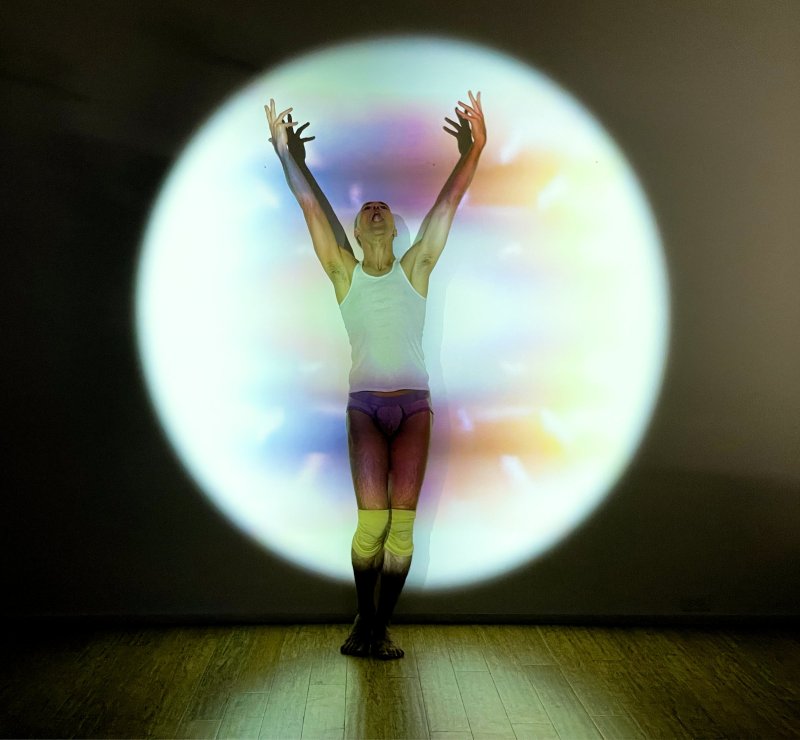 Performer in white tank top and purple briefs with arms raised, illuminated by a colorful circular projection on a dark wall.