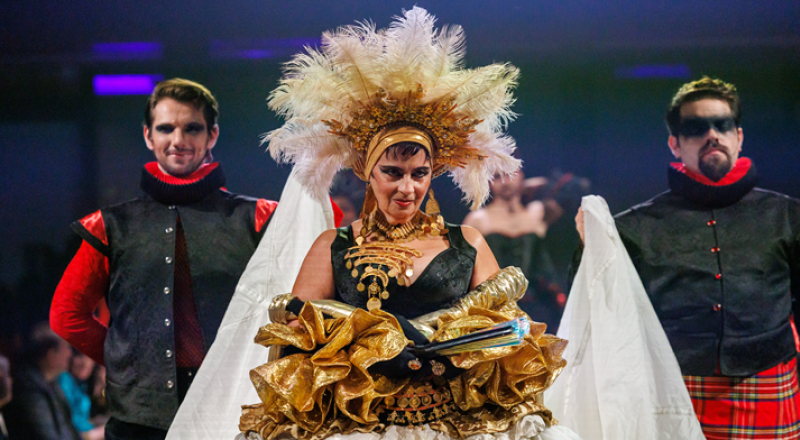 A performer in an elaborate gold and white feathered costume, flanked by two men in black and red outfits, on a runway.