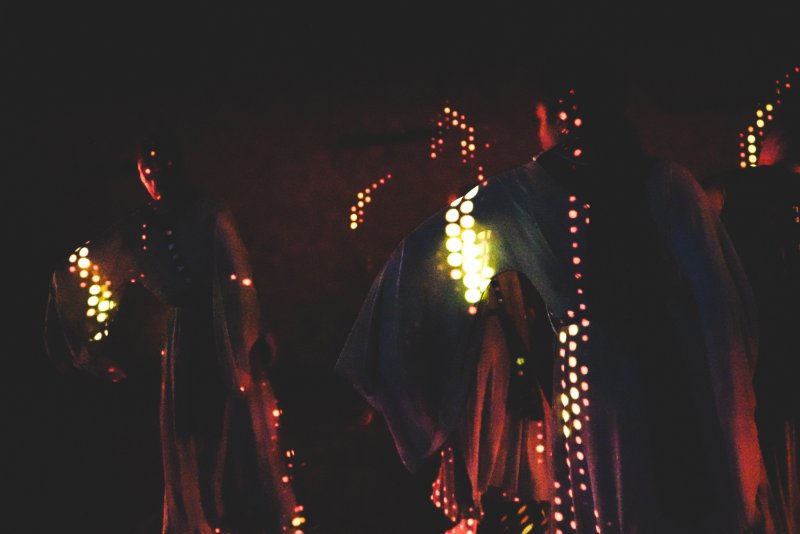 Two figures in dark, flowing robes are illuminated by vertical trails of glowing orange and yellow dots against a black background.