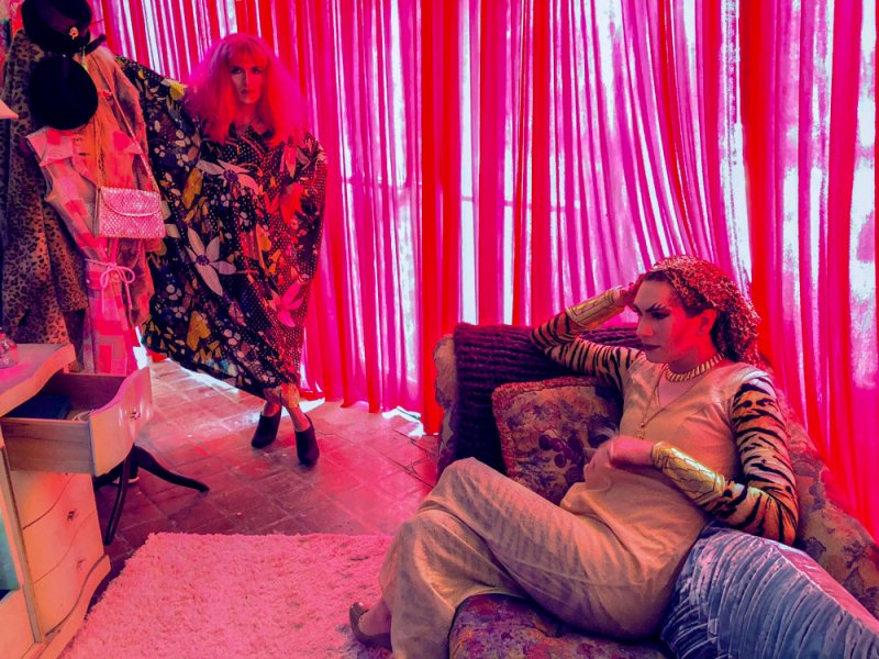 Two drag queens in a room with vibrant pink curtains. One stands in a floral robe, the other reclines in a tiger-print top.