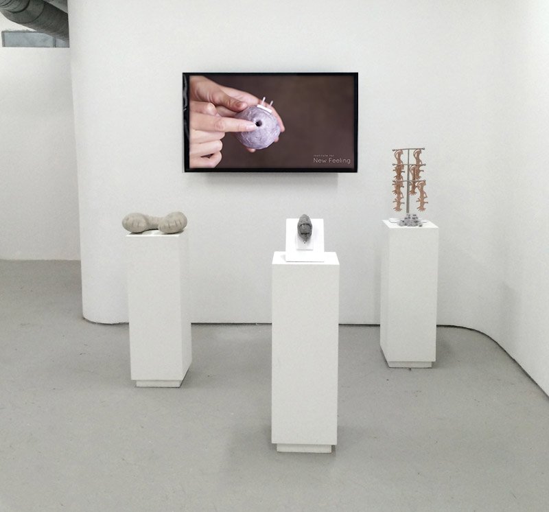 Gallery view with a video on a white wall and three white pedestals displaying small, abstract sculptures.