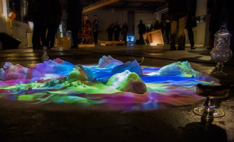 Installation with colorful light projected onto white mounds and scattered green spheres on a dark floor.
