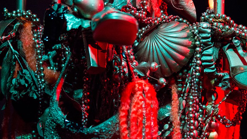Close-up of a dense sculpture with dark red and teal lighting, featuring beaded strands, shells, and shoes.