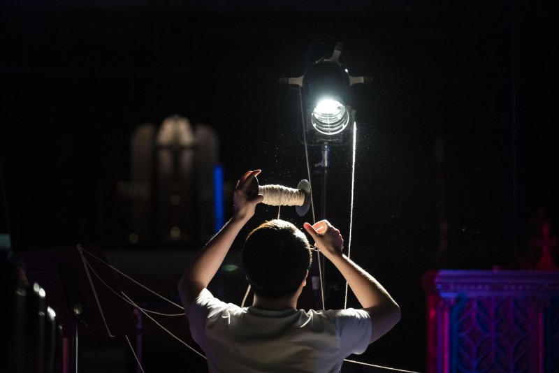 Person from behind, holding a spool of white string, illuminated by a bright stage light against a dark background.