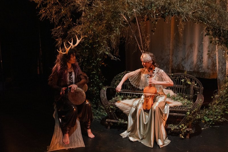 Two performers in costume, one with antlers playing a drum, the other with feathers playing a violin, in a forest-like setting.