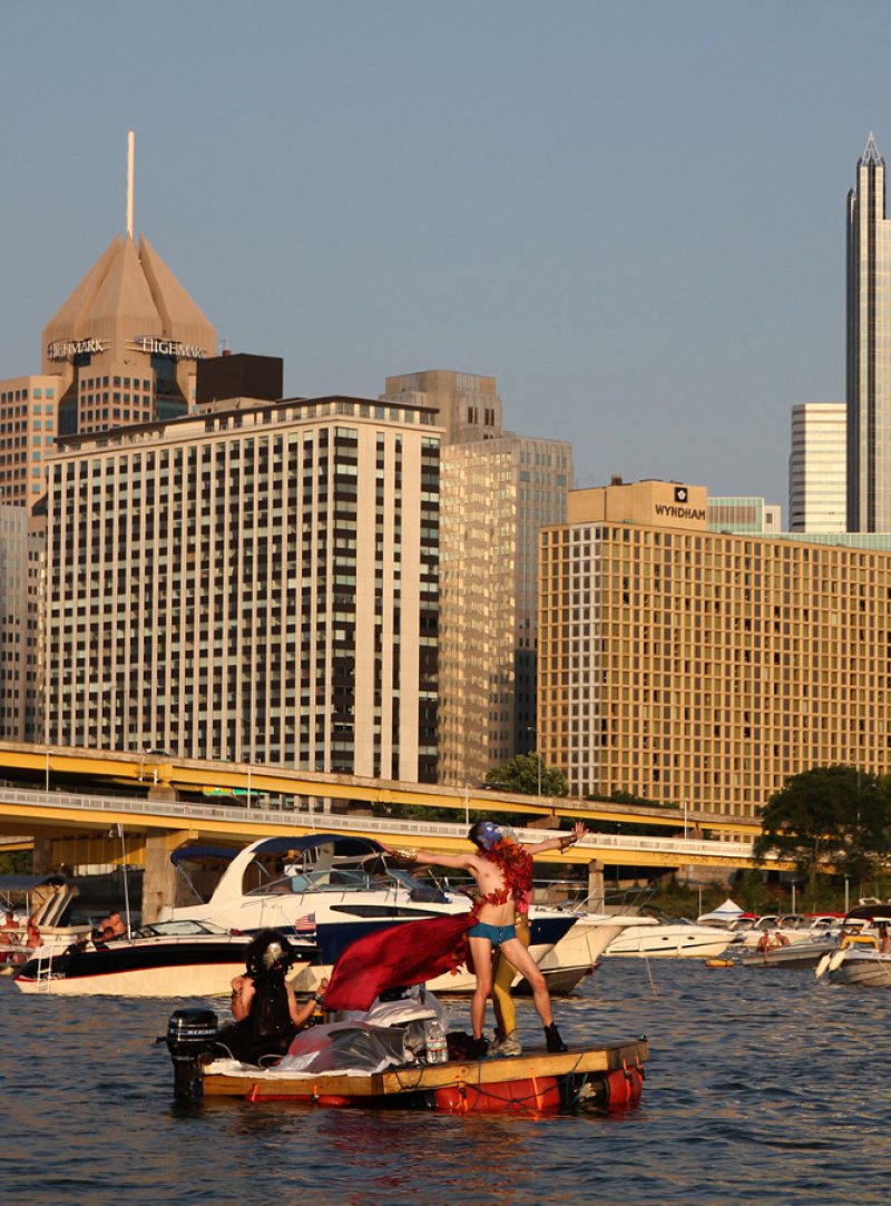 Two performers on a small raft in a river, with city buildings and boats in the background under a clear sky.