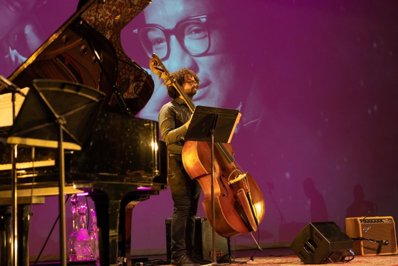 A musician with curly hair plays a cello on stage, a grand piano to his left, and a large purple projection of a man's face behind him.
