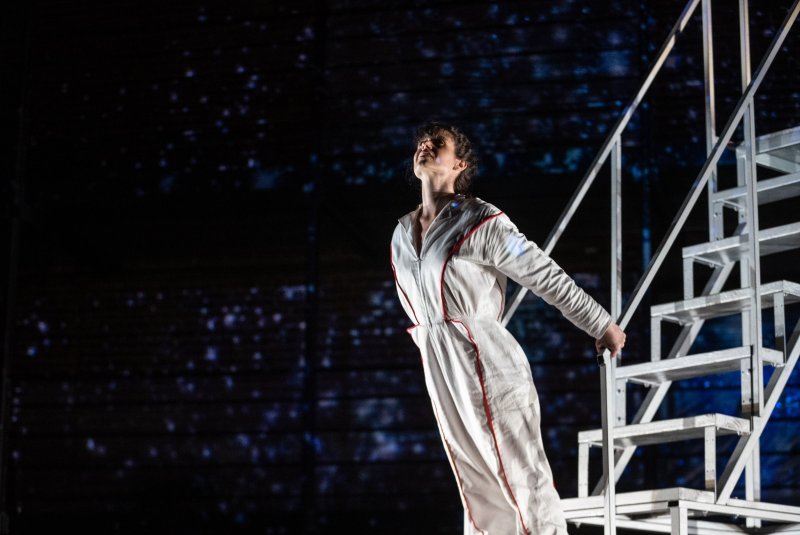 Woman in white jumpsuit with red trim, looking up, holding onto a metal staircase against a dark, starry background.