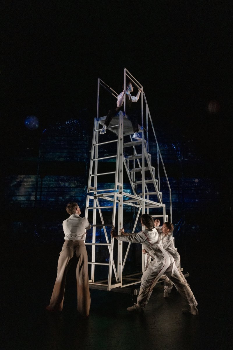 Four performers in white and tan costumes interact with a tall, white, geometric ladder structure on a dark stage with blue lighting.