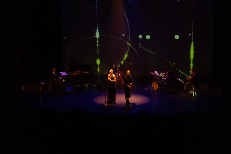 Two singers on a dark stage with musicians, illuminated by a spotlight, with green digital lines projected on the back wall.