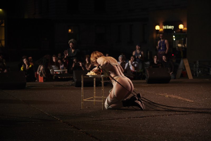 Performance art piece: Kneeling figure in lingerie eats from a table, cables extend behind. Audience blurred in background. Dark, outdoor setting.