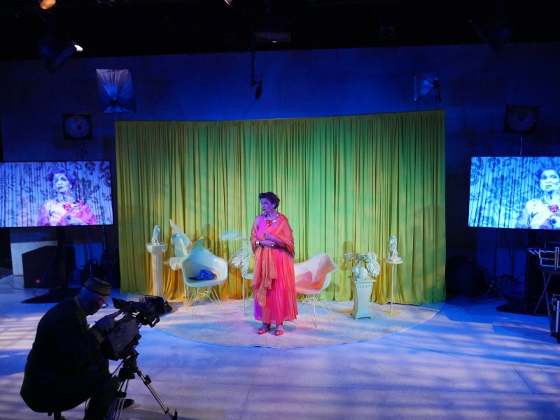 Performance art with a woman in an orange dress on a stage with yellow curtains, a cameraman, and two screens.