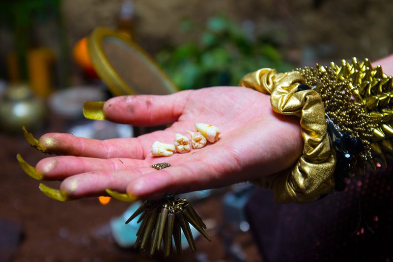 Hand with long golden nails and a spiked bracelet holds four small teeth and a tiny metallic beetle.