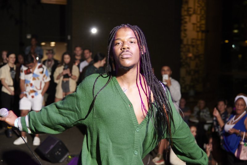 Performer with long braids, wearing a green cardigan, holds a microphone on stage. A blurred audience is visible in the background. The lighting is bright on the performer.