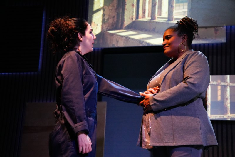 Two women, one in a dark jumpsuit, the other in a gray blazer, hold hands center stage with a projected backdrop of barred windows.