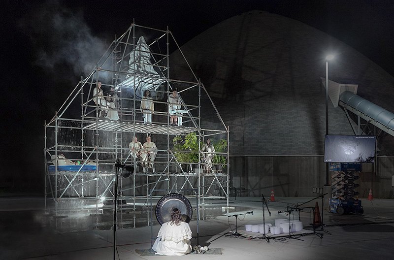 Performance art with people on a scaffolding structure under falling water, a gong, and a screen, against a dark, pyramid-like building.