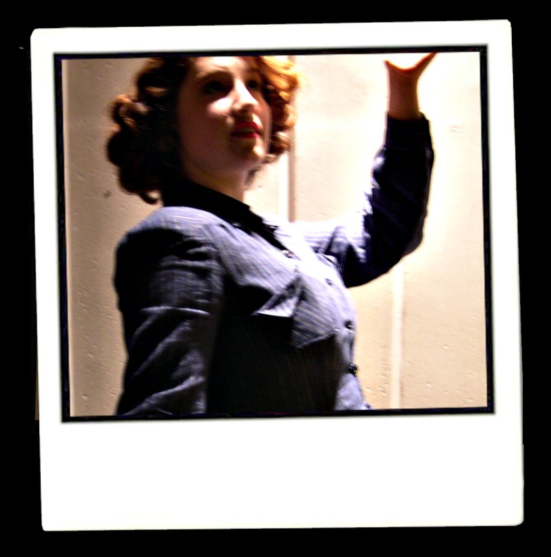 A woman with curly brown hair, wearing a dark pinstripe shirt, raises her left arm, bathed in warm light.
