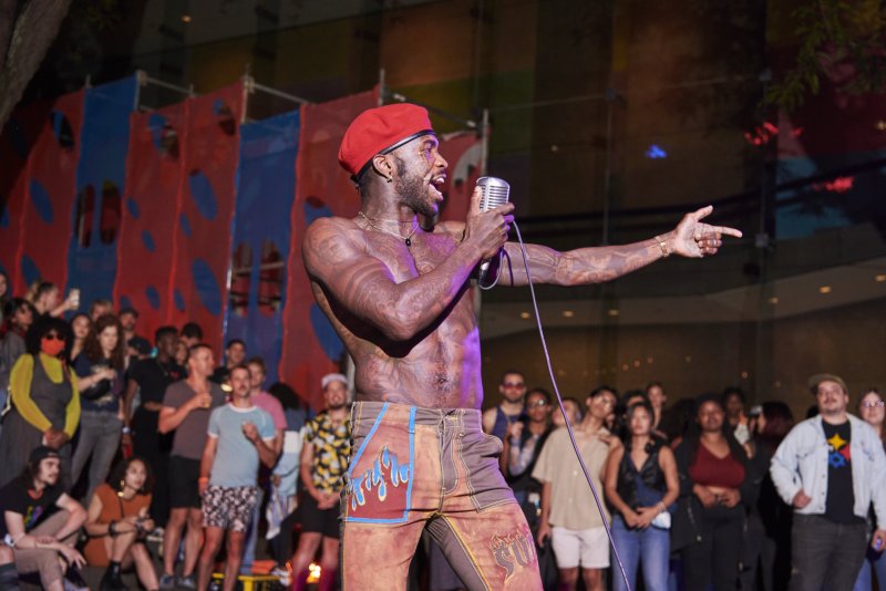 Performance art documentation: A shirtless, tattooed person in a red beret sings into a microphone on stage before a diverse crowd. They wear brown pants with blue flame designs. A red and blue backdrop is partially visible.