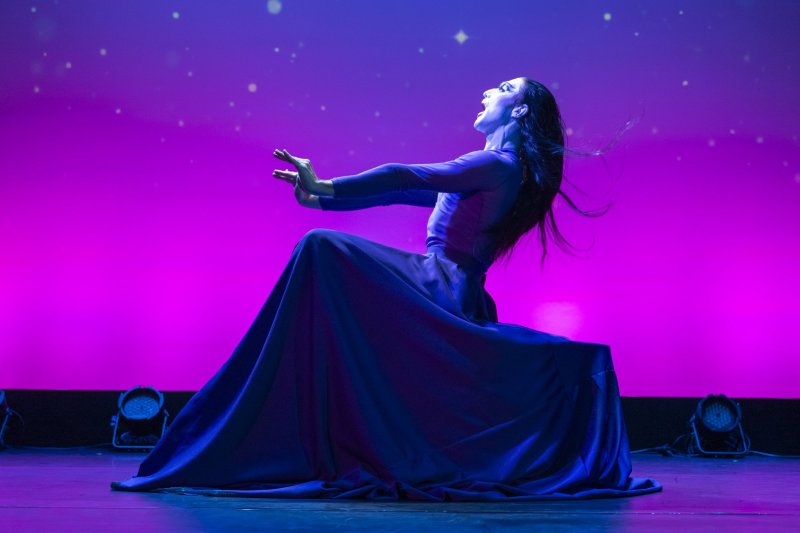 Dancer in blue gown, arms outstretched, head tilted back against a pink and purple starry backdrop.