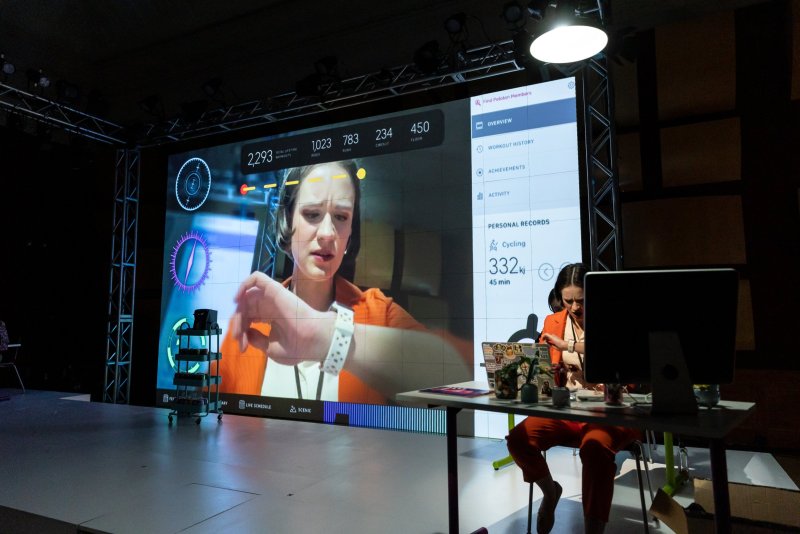 Installation view featuring a large screen displaying a woman's face and fitness data, juxtaposed with a seated figure in orange clothing at a desk with a computer. Stage lighting and metal framework visible.