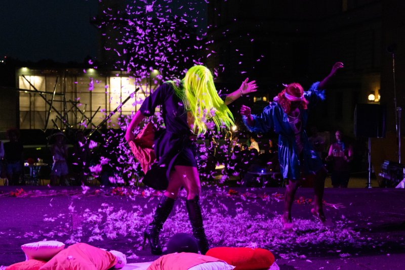 Two performers on a dark stage with purple lighting, one in a black dress and neon yellow wig, the other in blue, surrounded by falling purple feathers.