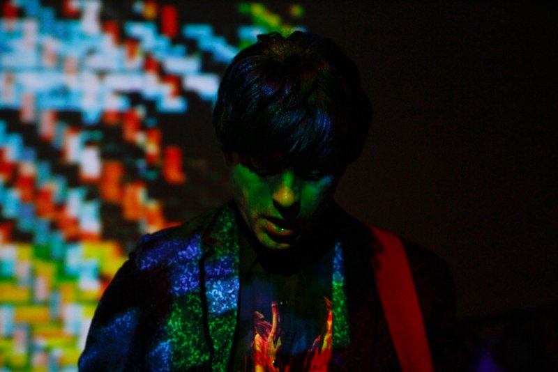 Person with green-lit face, patterned jacket, and flame shirt, against a pixelated background.