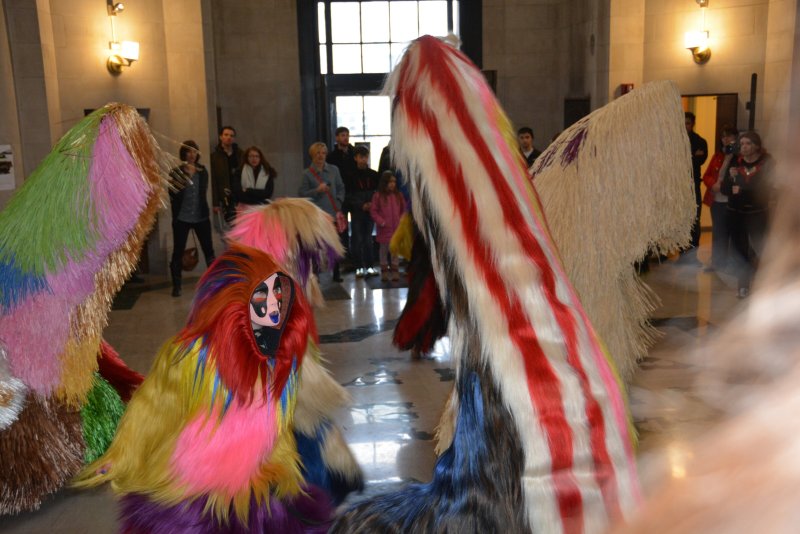 Performers in colorful, shaggy costumes with long tails dance in a large hall with high ceilings and windows.
