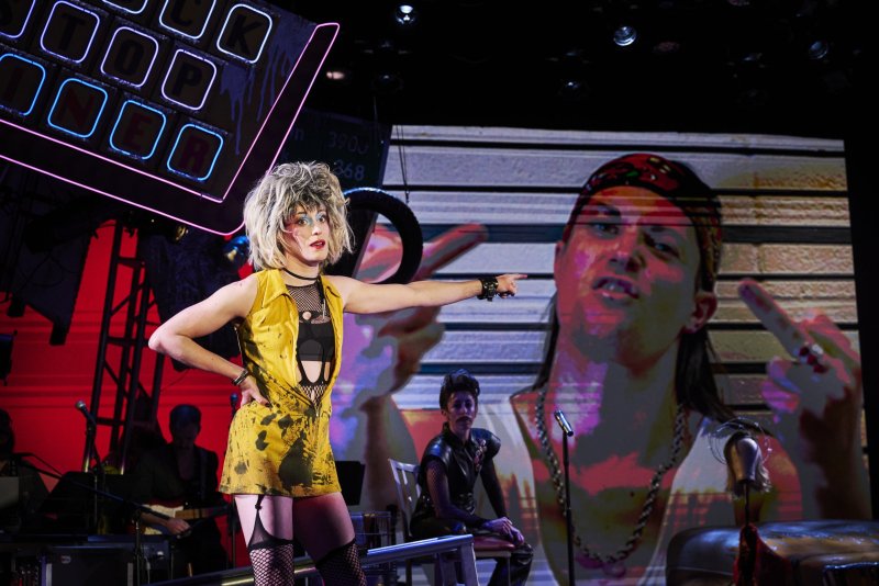 A blonde performer in a yellow dress and fishnets points right, a large screen behind them shows a person giving the middle finger.