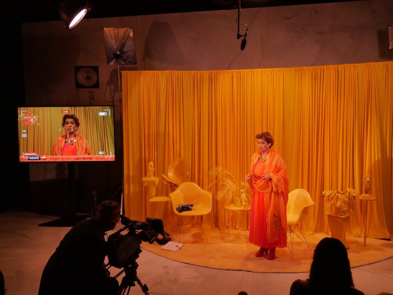 Woman in orange dress and shawl on a stage with yellow curtains, chairs, and objects, filmed by a camera operator.