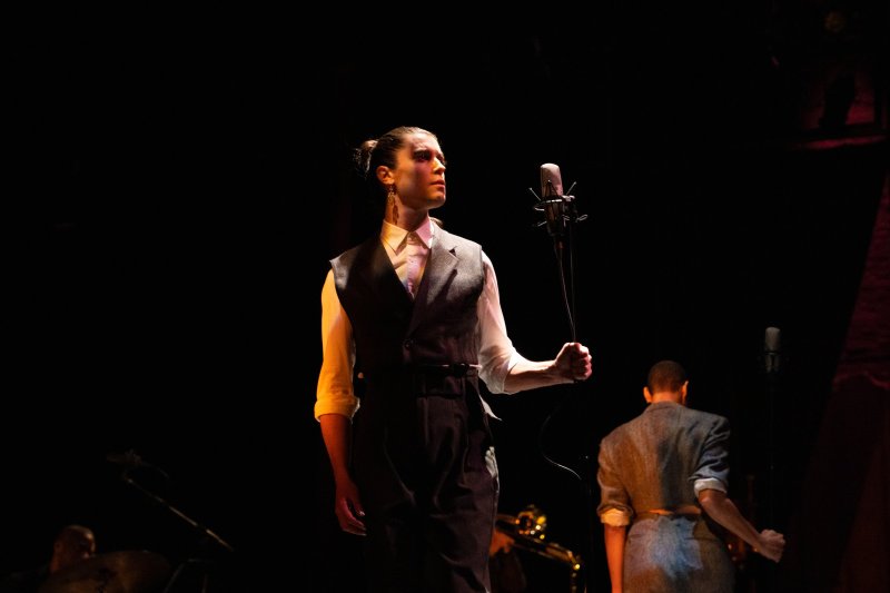 Performer in a dark vest and white shirt holds a microphone, looking right, with another performer's back visible in the foreground.