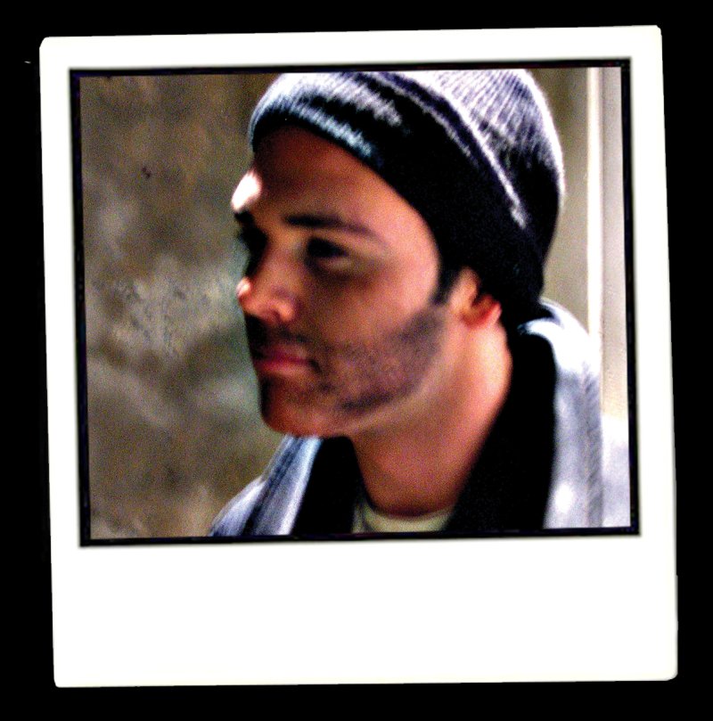 Polaroid of a man in a dark beanie and jacket, facing right, with smudged dark marks on his cheek and jaw, against a blurred background.