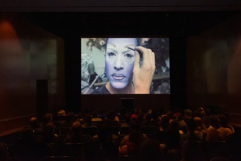 A drag queen with blue face paint and white eye makeup applies more makeup, projected on a large screen to an audience in a dark room.