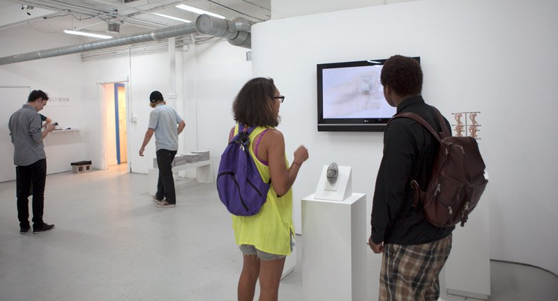 Two people viewing a screen and sculpture in a bright, minimalist gallery with other visitors and artwork.